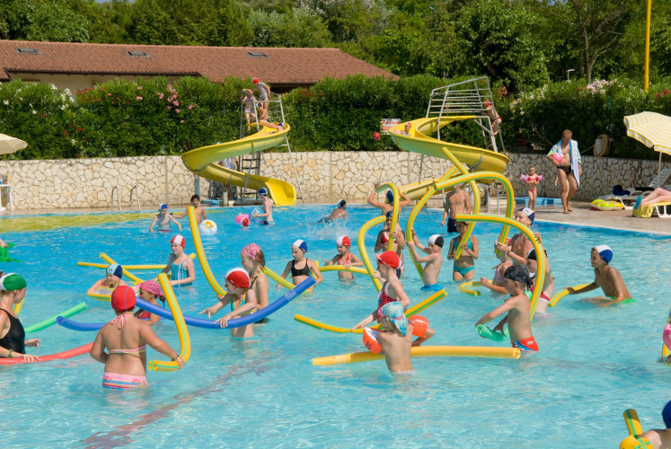 Children play with colorful pool noodles in a swimming pool with slides at a holiday park in Veneto, Italy.