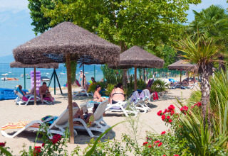 Persone rilassate su lettini sotto ombrelloni di paglia a Camping Cisano/San Vito, Veneto, Italia.