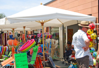 Outdoor market at Camping Cisano/San Vito, Veneto, Italy, with umbrellas, pool gear, and tourists browsing.