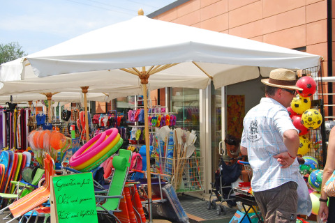 Marché extérieur au Camping Cisano/San Vito, Italie, avec parasols, bouées et vacanciers en promenade.