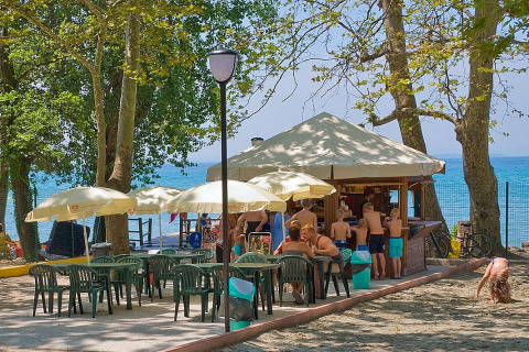 Cafetería al aire libre en Camping Cisano/San Vito con mesas, sombrillas y personas junto a la playa en Véneto, Italia.