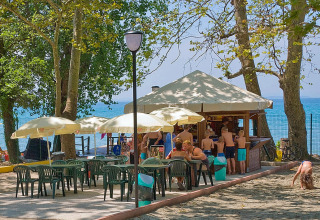 Udendørs café ved Camping Cisano/San Vito med borde, parasoller og folk, tæt på stranden i Veneto, Italien.