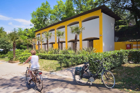 Una donna in bicicletta passa davanti a un edificio moderno con palme al Camping Cisano/San Vito, parco vacanze in Veneto.