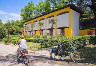 A woman rides a bicycle past a modern building with palm trees at Camping Cisano/San Vito holiday park in Veneto.