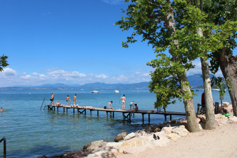 Camping Cisano/San Vito en Véneto, Italia, con muelle, bañistas y paisaje de lago bajo cielo azul.