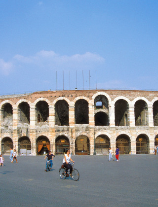 People walking and cycling in front of Verona Arena, a historic Roman amphitheater in Veneto, Italy.