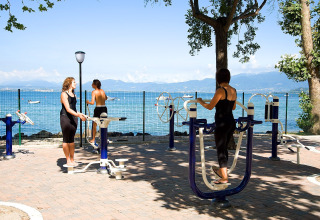 Outdoor gym at Camping Cisano/San Vito in Italy, people exercising on machines by a scenic lakeside view.
