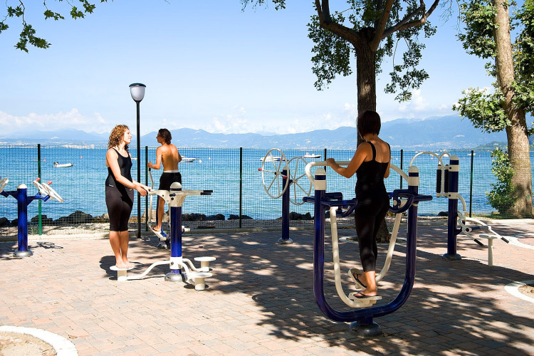 Outdoor gym at Camping Cisano/San Vito in Italy, people exercising on machines by a scenic lakeside view.