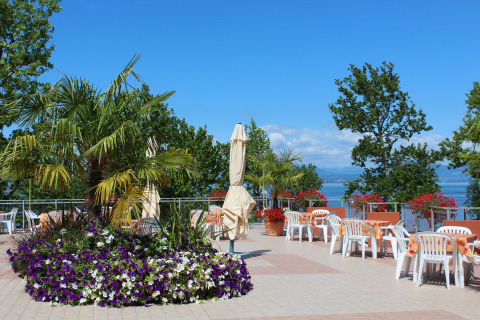 Terraza exterior con sillas y mesas blancas, flores y palmeras en Camping Cisano/San Vito, Véneto, Italia.