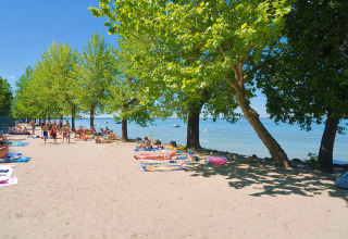 Beach area with sunbathers and trees at Camping Cisano/San Vito holiday park in Veneto, Italy, relaxing by water.