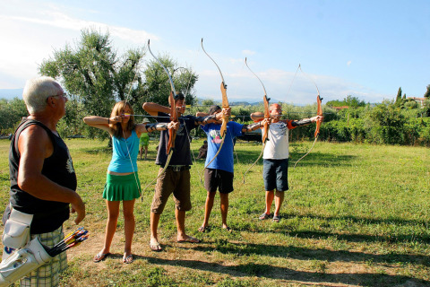 Four people practice archery on a sunny field while an instructor watches, at a holiday park in Italy.