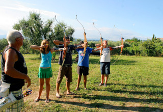 Four people practice archery on a sunny field while an instructor watches, at a holiday park in Italy.