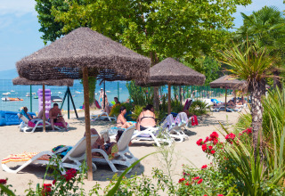 Persone che si rilassano su lettini sotto ombrelloni di paglia alla spiaggia del Camping Cisano/San Vito, Veneto.