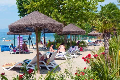 Vakantiegangers relaxen op ligstoelen onder rieten parasols op het strand van Camping Cisano/San Vito, Veneto.