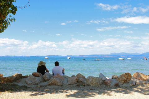 Two people sit on rocks by the lakeshore at Camping Cisano/San Vito, Veneto, Italy, overlooking the serene water.