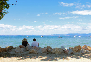 Two people sit on rocks by the lakeshore at Camping Cisano/San Vito, Veneto, Italy, overlooking the serene water.