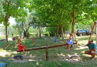 Two children play on a seesaw with an adult in a shady park surrounded by trees at Camping Cisano, Veneto, Italy.