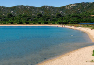 A peaceful sandy beach with clear blue water, people relaxing and swimming, green hills in the background.