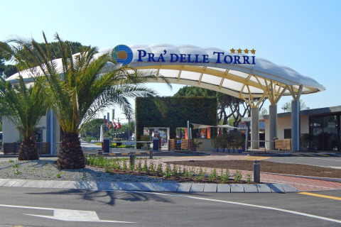 Entrada al Camping Pra'delle Torri en Véneto, Italia, con palmeras y un moderno arco en la entrada del parque.