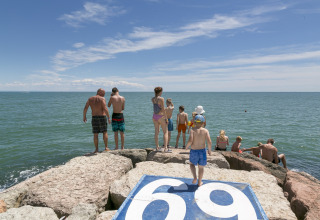 Familier nyder solen på store sten ved havet i Camping Pra'delle Torri, Veneto, Italien, med klart blå himmel.