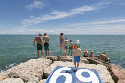 Familias disfrutan del sol sobre rocas junto al mar en Camping Pra'delle Torri, Véneto, Italia, bajo cielo azul.