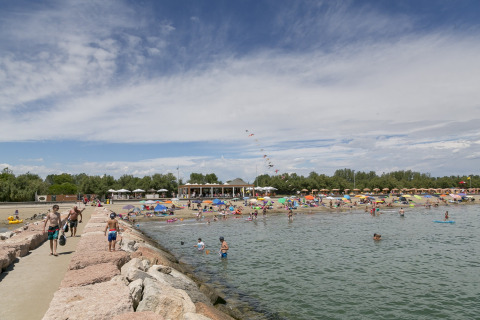 Escena de playa en Camping Pra'delle Torri en Véneto, Italia, con bañistas, sombrillas y cielo despejado.