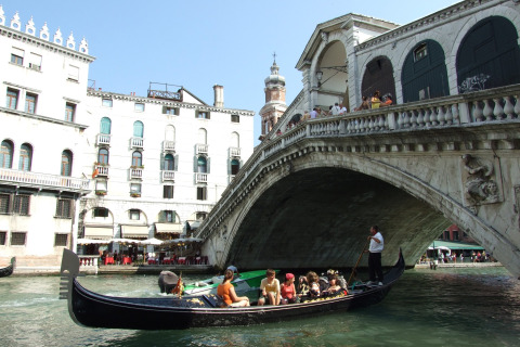 A gondola with tourists passes under the iconic Rialto Bridge in Venice, Italy, on a bright sunny day.