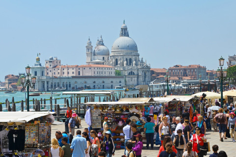 Bancarelle affollate e numerose persone davanti a una storica chiesa con cupola sul canale a Venezia, Italia.