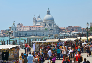 Travle markedsboder med mange mennesker foran en historisk kuppelbygning og kanal i Venedig, Italien.