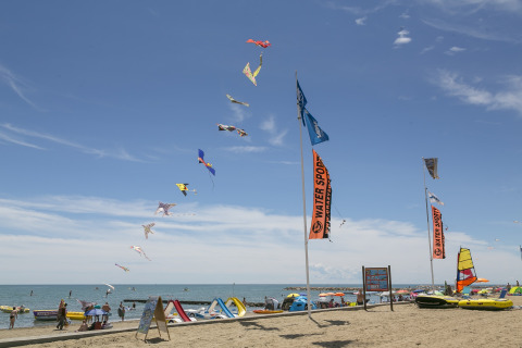 Escena de playa en Camping Pra'delle Torri, Veneto, Italia, con cometas coloridas y banderas de deportes acuáticos.