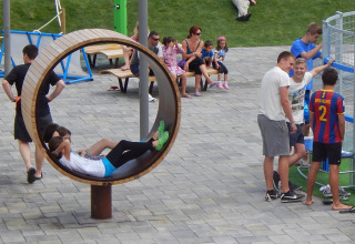 Børn leger i en cirkulær gynge i en park, mens voksne og teenagere nyder udendørsområdet ved Camping Pra'delle Torri.