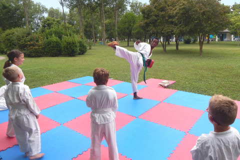 Niños aprenden artes marciales al aire libre mientras un instructor demuestra una patada en Camping Pra'delle Torri.