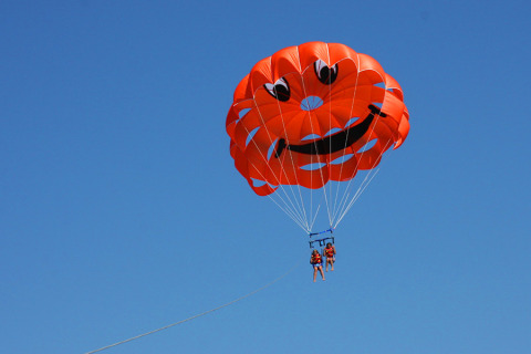 Two people parasailing with an orange parachute featuring a smiley face at Camping Pra'delle Torri, Italy.