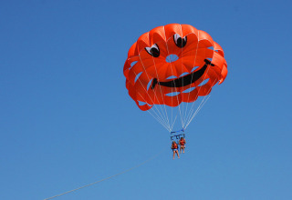 Two people parasailing with an orange parachute featuring a smiley face at Camping Pra'delle Torri, Italy.