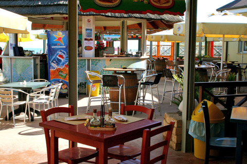 Cafetería al aire libre en Camping Pra'delle Torri, Véneto, Italia, con mesas, sillas y sombrillas visibles.