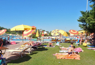 Holiday park guests relax on sunbeds and grass under colorful umbrellas by the pool at Camping Pra'delle Torri.