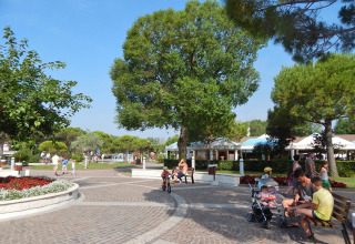 Sunny plaza at Camping Pra'delle Torri, Veneto, Italy, with families enjoying lush trees and flower beds.