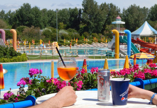 To personer nyder drinks ved et poolområde med vandrutsjebaner og parasoller i Camping Pra'delle Torri, Italien.