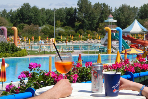 To personer nyder drinks ved et poolområde med vandrutsjebaner og parasoller i Camping Pra'delle Torri, Italien.