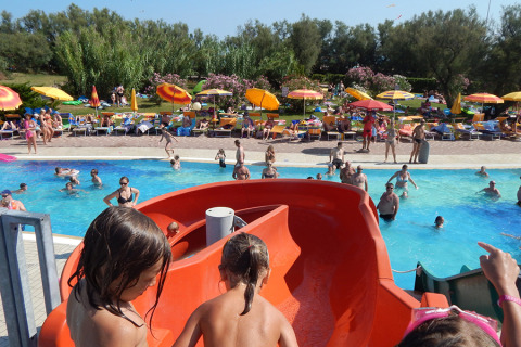 Niños listos para deslizarse por un tobogán rojo en la piscina concurrida de Camping Pra'delle Torri, Italia.