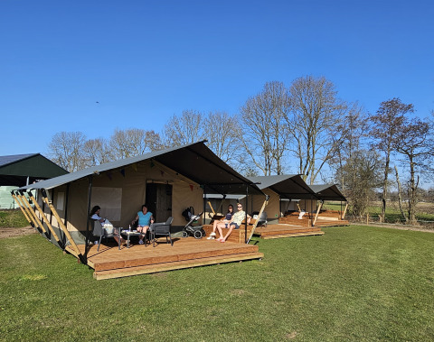 Tenda safari con bagno, persone rilassate sulla veranda sotto un cielo azzurro circondate da alberi.