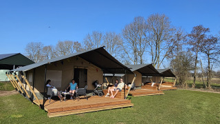 Safaritent met toilet en mensen relaxen buiten op een houten terras onder een heldere blauwe hemel.