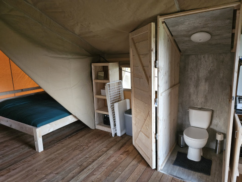 Interior of a safari tent with wooden floor, bed, shelving, and private toilet at HappyFarm in the Netherlands.