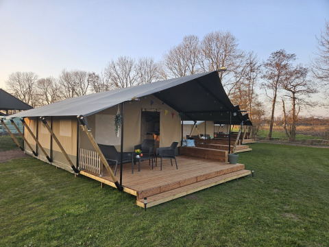 Safaritent met houten terras en zitje bij The HappyFarm in Nederland, omgeven door gras en bomen.