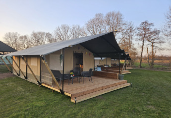 Tenda safari con veranda in legno e posti a sedere a The HappyFarm nei Paesi Bassi, tra prato e alberi.