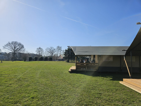 Safari telt med toilet på The HappyFarm i Holland, med græsplæne, solskin og folk på terrassen.