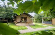 Tienda glamping familiar en Urban-Gardens Ieper, Bélgica, rodeada de césped verde y frondosos árboles.