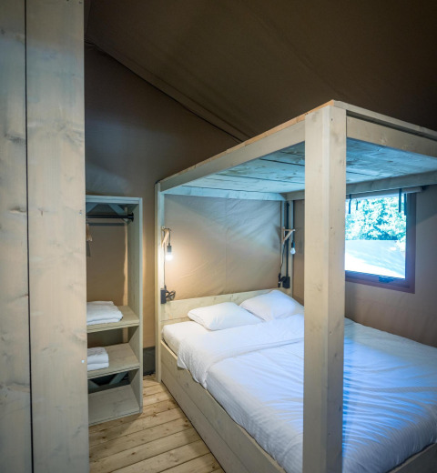 Interior de tienda safari con cama doble, suelo de madera, estantería abierta y luz natural por la ventana.