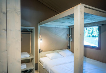 Interior of a safari tent with a canopy bed, wooden floor, open shelving unit, and bright window view.