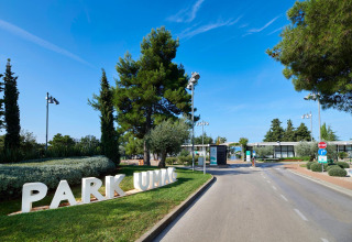 Indgangen til Camping Park Umag i Istrien, Kroatien, omgivet af grønne træer og blå himmel.